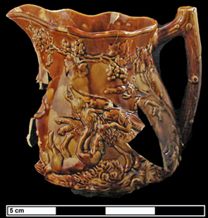 Side view of a molded yellow and brown pitcher with a scene of dogs hunting a wild pig.